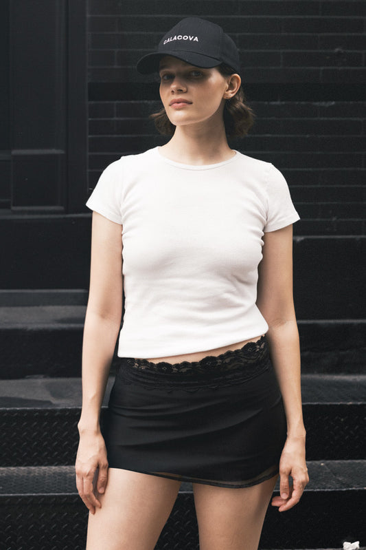 Woman wearing a white t-shirt and black skirt with a cap against a black brick wall.